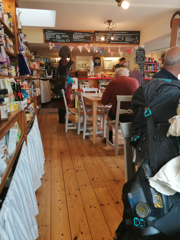 Interior of a small cafe or shop with wooden floors. A Rastafarian in a long dark brown coat and hat is near the counter assisting customers. Several people are seated at light gray tables, eating and drinking. Shelves stocked with various goods line one wall. The atmosphere is quaint and rustic. A large backpack is prominently displayed in the foreground, partially obscuring some seated customers.