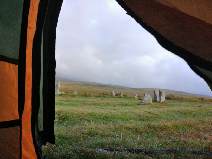 View from inside a tent, looking out at a field with several standing stones. The stones appear to be part of a stone circle or similar ancient monument. The sky is overcast. The overall impression is one of peaceful solitude in a wild, somewhat remote location.