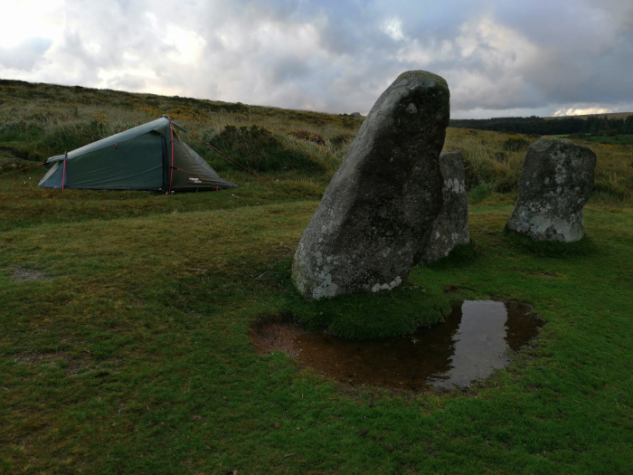 Solitary green tent pitched on a grassy hill, near a cluster of large, weathered stones that resemble a small, incomplete stone circle.  A small puddle of water is visible near the stones. The sky is overcast. The overall impression is one of peaceful solitude in a remote, possibly ancient, location.