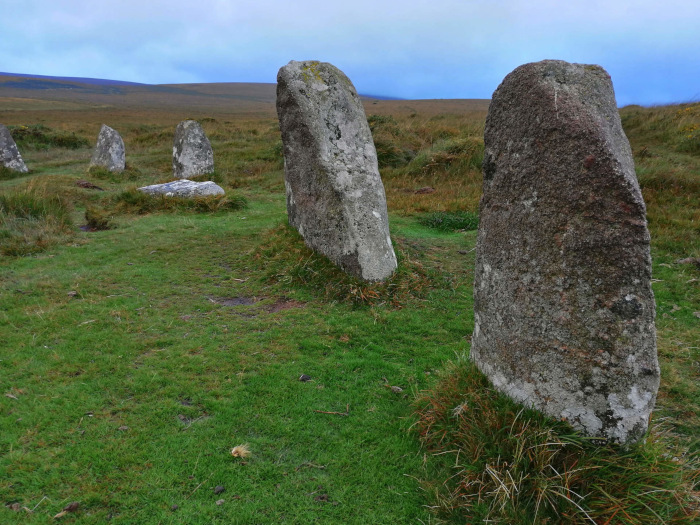Stone circle, a type of megalithic monument, set in a grassy field under a cloudy sky. Several large, upright stones are visible, some more weathered than others, with one prominent stone in the foreground. The scene evokes a sense of age and history.