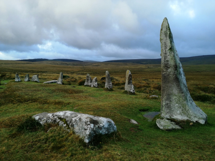 Stone circle, a type of megalithic monument, set in a grassy moorland under a cloudy sky. The stones vary in height and shape, with one particularly tall and slender stone dominating the right side of the frame. The overall atmosphere is one of antiquity and quiet solitude.