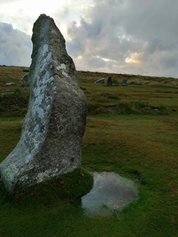 Single, large, grey standing stone in a grassy field under a cloudy sky. A small puddle of water is at the base of the stone. In the background, other, smaller standing stones are visible, suggesting a stone circle or similar ancient monument. The overall impression is one of quiet solitude and the passage of time.