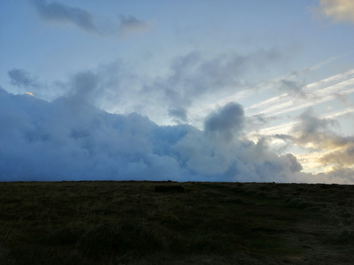 Vast, flat expanse of dark brown moorland under a dramatic sky. A thick, rolling layer of clouds dominates the upper two-thirds of the frame, creating a moody, almost ominous atmosphere. The clouds are a blend of dark blue and gray, with hints of lighter colour suggesting either the setting or rising sun. The lower third is a relatively dark, unblemished field, suggesting a desolate or remote location. The overall effect is one of brooding stillness and expansive space.