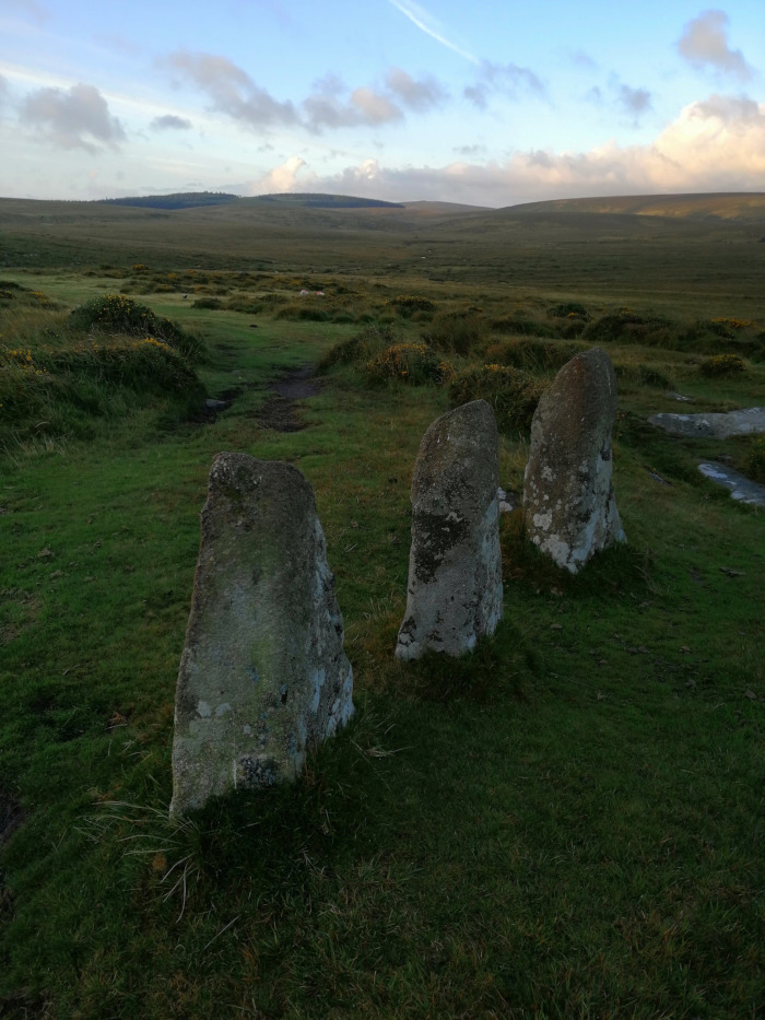 Three upright stones, possibly part of a stone circle or other ancient structure, standing in a grassy field under a cloudy sky. The background is a vast, rolling moorland. The scene evokes a sense of antiquity and remoteness.