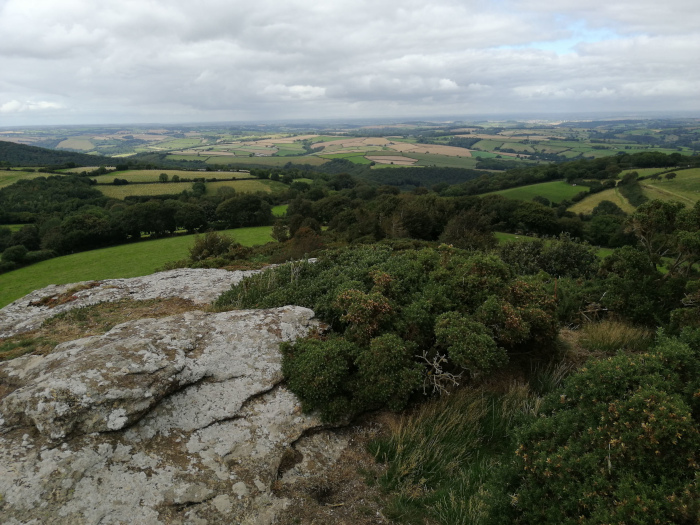 Panoramic view of a rolling green landscape, likely a rural countryside, seen from an elevated vantage point, possibly a hilltop. The foreground features a large, lichen-covered rock formation, with low-lying shrubs and grasses surrounding it. The middle ground shows a dense forest or woodland descending into more open pastureland. The background is a wide expanse of fields and valleys extending to the horizon under a slightly overcast sky.