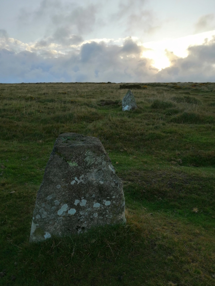 Wide, open grassy field under a cloudy sky. Two large, weathered stones are prominently featured in the foreground and mid-ground. The stones appear old and possibly part of a historical site or natural formation. The overall mood is serene and somewhat melancholic, with a sense of vastness and antiquity. The setting sun is visible through the clouds in the background.