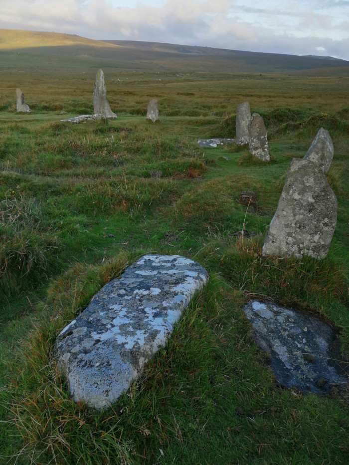 Stone circle, likely of ancient origin, situated in a grassy field. Several upright stones of varying heights and shapes are partially visible, some fallen or broken. One large, flat stone lies on the ground, resembling a slab or capstone. The setting is rural and seemingly remote, with a low hill visible in the background under a somewhat cloudy sky. The overall impression is one of age, history, and the passage of time.