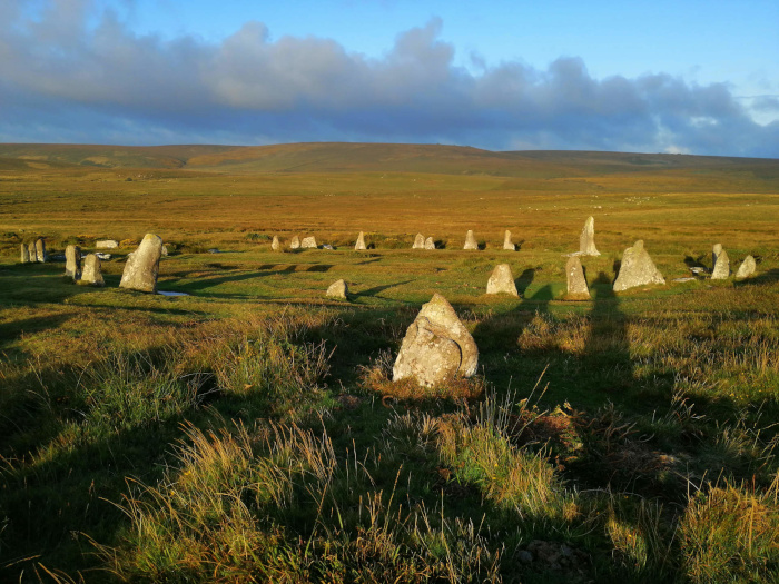 Stone circle in a grassy field, possibly in Scotland or Ireland. The stones are various heights and shapes. The setting sun casts long shadows across the field. The overall feeling is one of quiet antiquity and the vastness of the landscape. The sky is partly cloudy with a hint of blue.