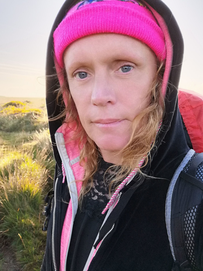 Close-up of Leonie's face. She appears to be outdoors, in a grassy field, wearing a black hooded jacket, a bright pink beanie, and a backpack. Her expression is serious, almost pensive. The lighting suggests it might be sunrise or sunset. The overall mood is contemplative and slightly melancholic.