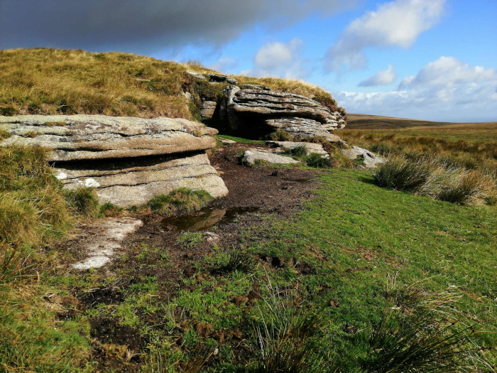 Natural rock formation, possibly a small cave or overhang, nestled into a grassy hillside. The rocks are layered and appear weathered, showing signs of erosion. The surrounding landscape is open and consists of short grass and patches of taller vegetation. The sky is partly cloudy, with blue sky visible in between the clouds. The overall impression is one of a tranquil, natural scene, possibly in a moorland or upland area.