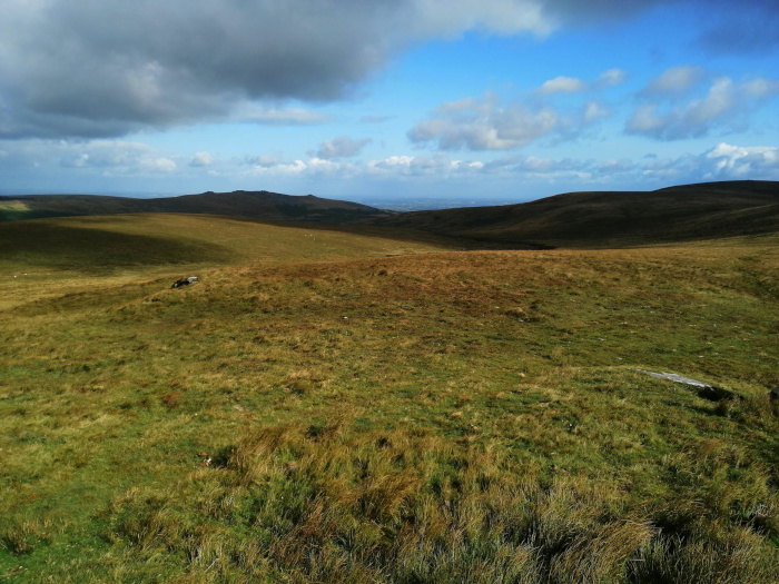 Vast, rolling landscape of grassy hills under a partly cloudy sky. The foreground is dominated by shorter, greener grass, while the hills in the middle and background show a mix of greens and browns, suggesting different types of vegetation or varying levels of sun exposure. The overall feeling is one of wide-open space and natural beauty, characteristic of a moorland or upland area. A hint of distant lowlands is visible on the horizon.