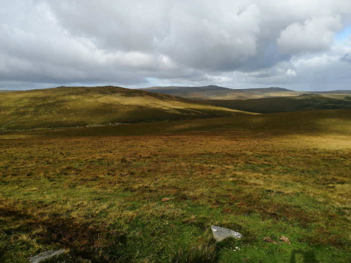 Vast, undulating expanse of moorland under a partly cloudy sky. The foreground is a field of low-lying vegetation, predominantly various shades of brown and green. In the mid-ground and background, softly rolling hills extend to the horizon, suggesting a remote and possibly mountainous region. The overall impression is one of wide open space and natural beauty, possibly a scene from a highland or moorland area.