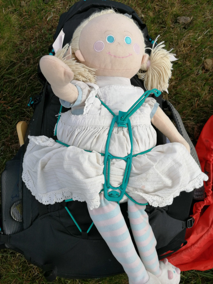 Rosie rag doll, dressed in a white dress and pink and white striped tights, strapped to the outside of a black backpack using a teal-coloured plastic harness. The backpack is sitting on a patch of grass. The doll appears well-loved, with some visible wear and tear on her dress.