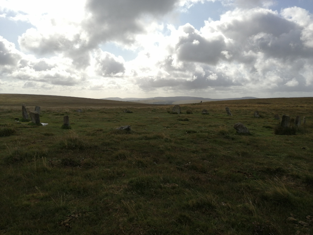 Wide shot of a grassy field under a cloudy sky. Scattered across the field are a number of upright stones, appearing to form a rough circle or ring. The overall impression is one of a remote, ancient, and possibly ceremonial site, possibly a stone circle. The muted colors and cloudy sky contribute to a somewhat melancholic or contemplative mood.