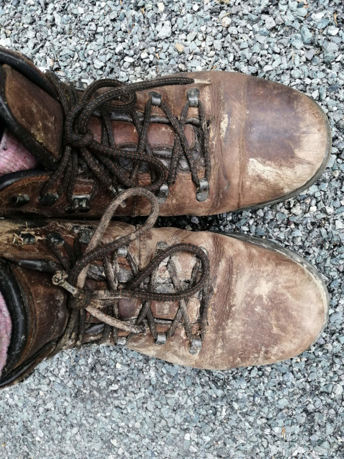 Pair of well-worn brown leather hiking boots, laced up and resting on a gravel surface. The boots are heavily scuffed and show significant signs of use, with dirt and wear visible across the leather. The laces are dark brown or black. The background is a bed of small, grey stones.