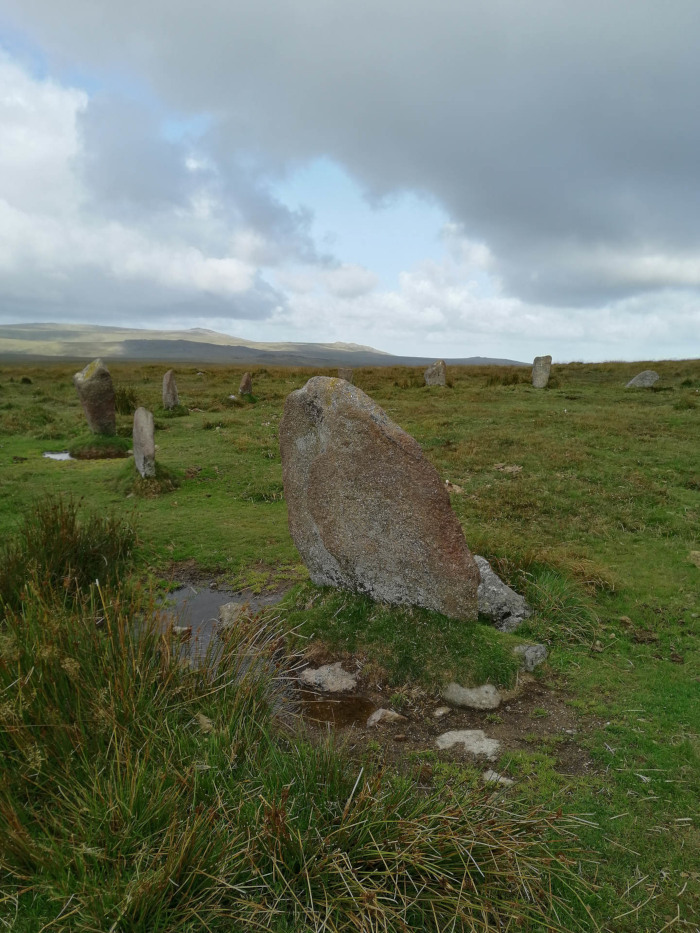 Stone circle, specifically what appears to be the remains of a prehistoric monument. Several large, upright stones are partially visible, set in a grassy field under a somewhat cloudy sky. The stones are irregular in shape and size, and some appear to be partially buried or fallen. The landscape is open and relatively flat, with low-lying vegetation. A few small puddles of water are visible in the foreground.