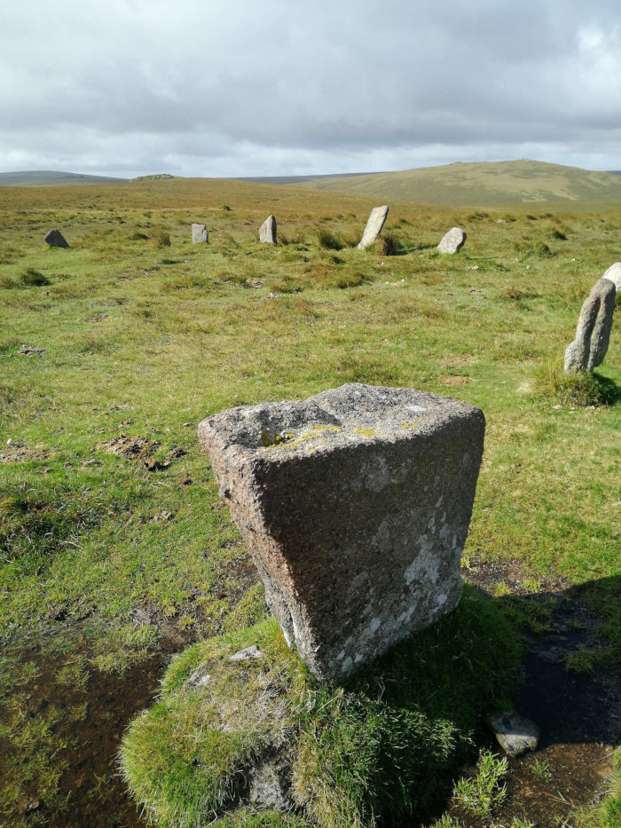 Stone structure, possibly a part of a larger stone circle or monument, situated in a grassy field under a cloudy sky. The foreground features a large, weathered, cube-like stone. In the background, several other upright stones are visible, suggesting the remains of an ancient site.