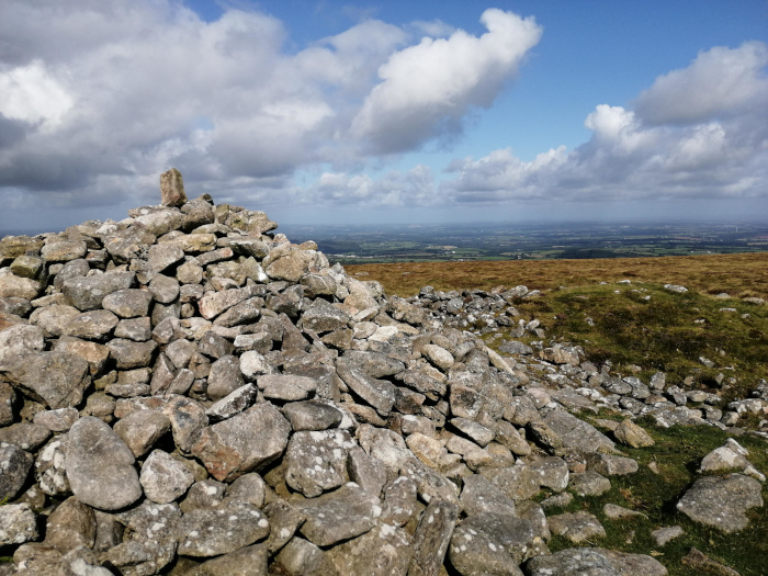 Cairn, a man-made pile of stones, situated on a hilltop. The cairn is composed of many grey and brown rocks of varying sizes, with a slightly larger, lighter-coloured stone at its apex. Beyond the cairn, the landscape extends into a distant view of rolling hills and fields under a partly cloudy sky. 