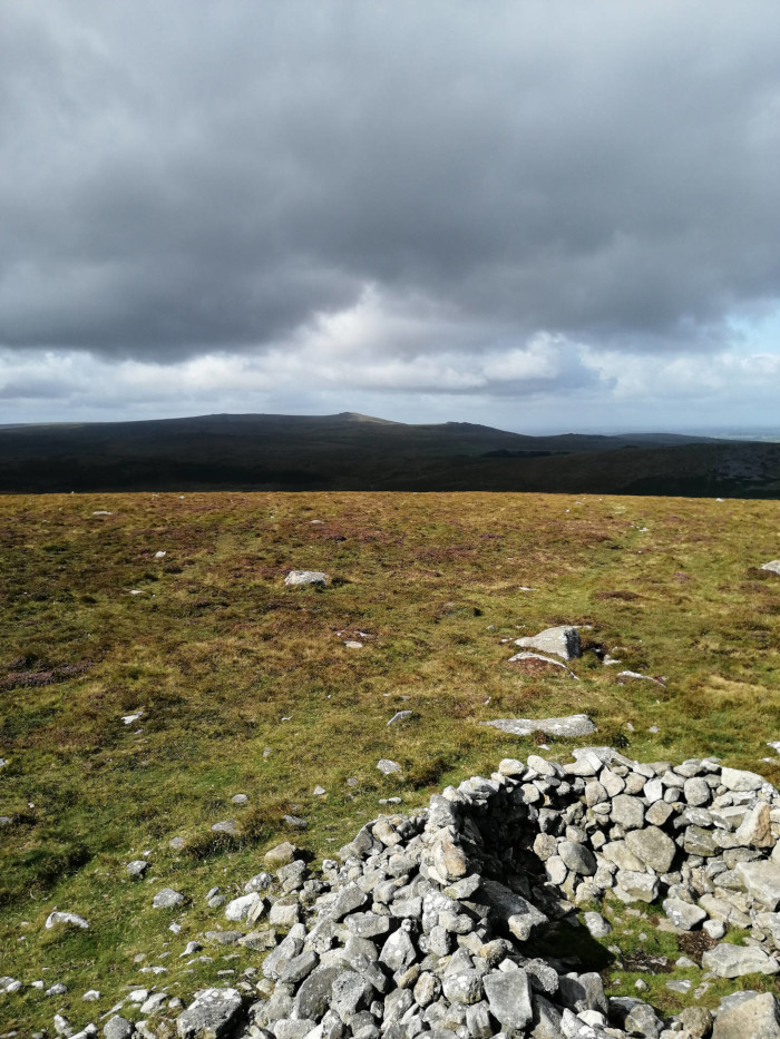 Bleak, windswept landscape under a brooding sky.  In the foreground is a low stone structure, possibly a ruin or ancient wall, partially overgrown with grass. The midground is a vast expanse of low-lying, sparsely vegetated land. In the distant background, a low hill or range of hills is visible under a sky filled with dark, heavy clouds that suggest an impending storm or unsettled weather.