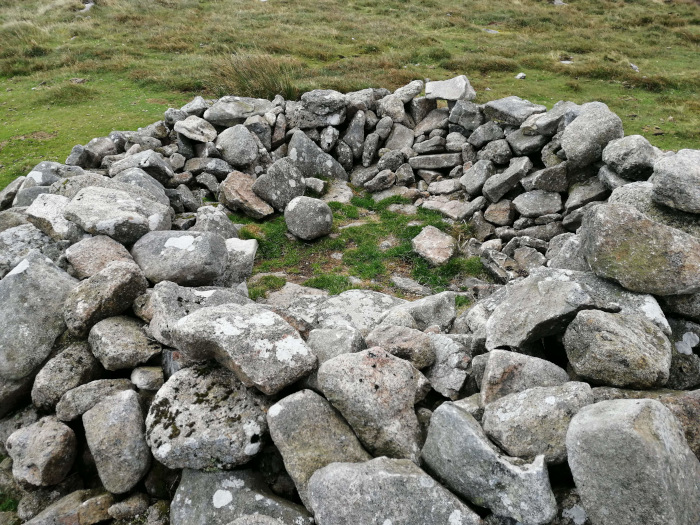 Circular stone structure, possibly a ruin or a low, dry-stone wall, situated in a grassy field. The stones are grey and irregularly shaped, varying in size. The interior of the structure is partially filled with grass. The overall impression is one of age and possible historical significance, suggesting a remnant of a past structure or boundary.