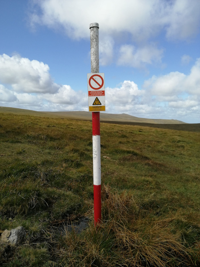 Red and white striped post in a grassy field under a partly cloudy sky. The post has a warning sign attached, indicating danger and prohibiting entry. The sign suggests the presence of unexploded ordnance or other hazardous materials. The overall scene is desolate and emphasises the warning, highlighting the potential danger in the area.