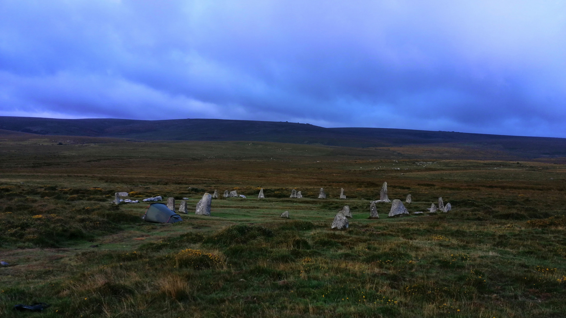 A lone tent pitched in a grassy field near a ring of standing stones under a twilight sky. The scene evokes a sense of solitude and connection to ancient history. The minimalist composition emphasises the vastness of the landscape and the smallness of the human presence within it.