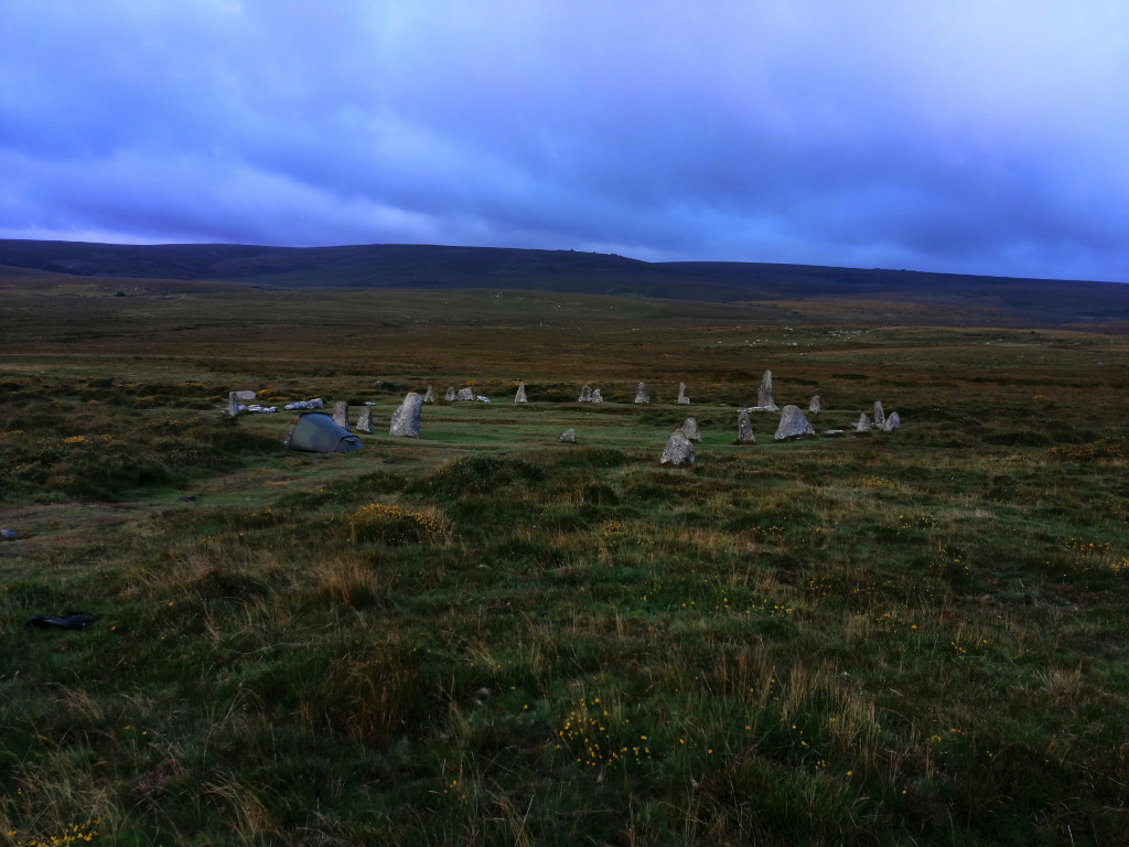 Lone tent pitched in a grassy field near a ring of standing stones under a twilight sky. The scene evokes a sense of solitude and connection to ancient history. The minimalist composition emphasises the vastness of the landscape and the smallness of the human presence within it.