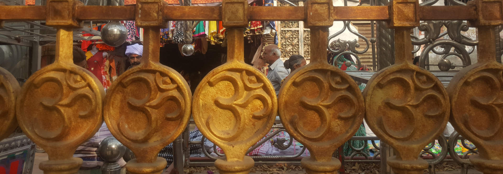 Rusty-gold metal fence, its vertical bars adorned with circular medallions bearing the Om symbol, a sacred Hindu sign. The fence acts as a visual barrier, partially obscuring the scene behind it. Behind the fence, several individuals are visible, appearing to be in a marketplace or shop.