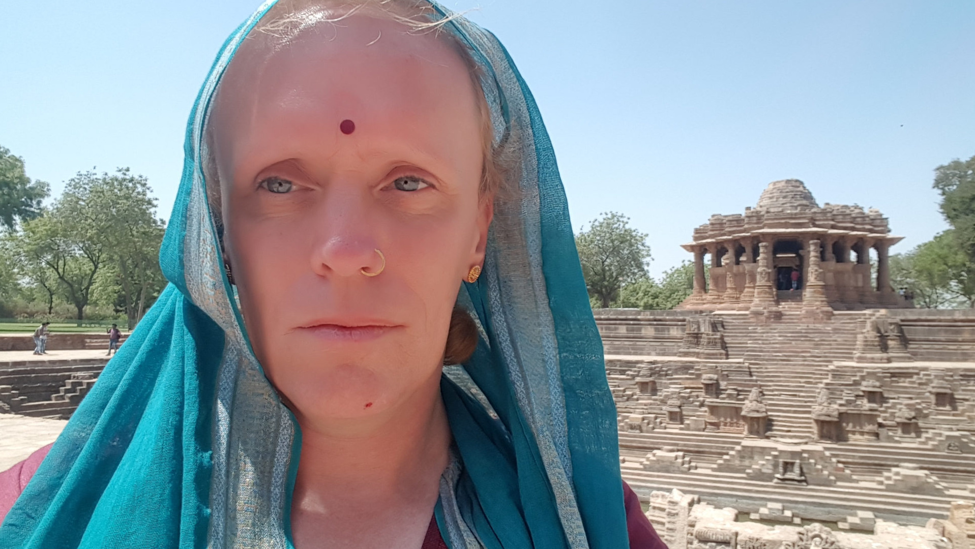 Leonie with short, light blonde hair partially visible under a teal and silver-striped sari or shawl. The sari is draped over her head and shoulders, covering most of her hair and framing her face. She's wearing a burgundy-coloured garment underneath, slightly visible at the neckline. A small, dark red bindi adorns her forehead, and a small gold nose ring is visible in her left nostril. The person is positioned slightly off-centre, looking directly at the camera, with a neutral to slightly serious expression. Their gaze is direct and steady, creating a sense of intimacy. The background is a sun-drenched, ancient-looking stepped well or water reservoir, a common feature in Indian architecture. A small, intricately carved sandstone temple-like structure sits atop the steps of the well. The well's architecture is complex, showcasing many levels of steps and detailed carvings. The surroundings include sparse trees and a clear blue sky. The overall lighting suggests it's daytime, with bright sunlight illuminating the scene. The color palette is warm and earthy. The teal of the sari provides a strong contrast to the muted browns and oranges of the stone architecture and the background. The sky is a clear, almost cloudless, vibrant blue.