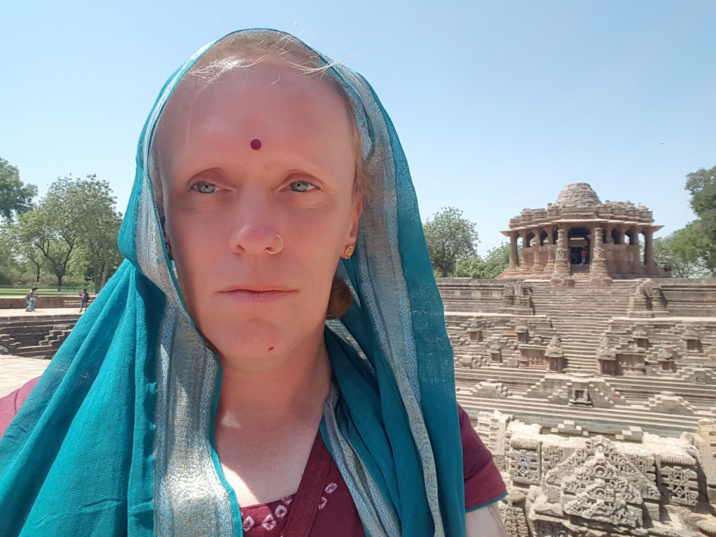 Leonie with short, light blonde hair partially visible under a teal and silver-striped sari or shawl. The sari is draped over her head and shoulders, covering most of her hair and framing her face. She's wearing a burgundy-coloured garment underneath, slightly visible at the neckline. A small, dark red bindi adorns her forehead, and a small gold nose ring is visible in her left nostril. The person is positioned slightly off-centre, looking directly at the camera, with a neutral to slightly serious expression. Their gaze is direct and steady, creating a sense of intimacy. The background is a sun-drenched, ancient-looking stepped well or water reservoir, a common feature in Indian architecture. A small, intricately carved sandstone temple-like structure sits atop the steps of the well. The well's architecture is complex, showcasing many levels of steps and detailed carvings. The surroundings include sparse trees and a clear blue sky. The overall lighting suggests it's daytime, with bright sunlight illuminating the scene. The color palette is warm and earthy. The teal of the sari provides a strong contrast to the muted browns and oranges of the stone architecture and the background. The sky is a clear, almost cloudless, vibrant blue.