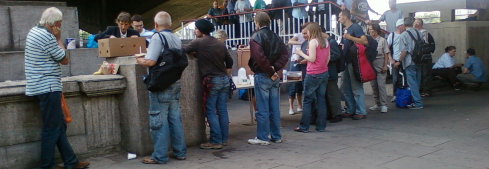 Group of people gathered under a bridge, seemingly interacting casually. A man with short, white hair, wearing a horizontally striped blue and white shirt and dark trousers, stands prominently on the left, slightly turned away, his hand near his face as if in thought. He stands near a low stone wall. Another man, wearing faded blue jeans and a dark jacket, is centrally positioned, holding a cardboard box. He has short, light brown hair. Next to him, a tall man with short, brown hair in a leather jacket stands with arms crossed. A collection of individuals is positioned behind and around the central figures. Many are wearing everyday clothing – jeans, jackets, t-shirts in muted colours; some with backpacks. Among them, a woman in a bright pink tank top and denim jeans stands out. A young man with dark hair and a brown jacket is further back, leaning slightly. A group of figures are visible above and slightly behind the main group, standing on a walkway/platform bordered by a railing. They are less clearly defined and appear to be watching or waiting. A higher structure suggests the image takes place under a bridge.