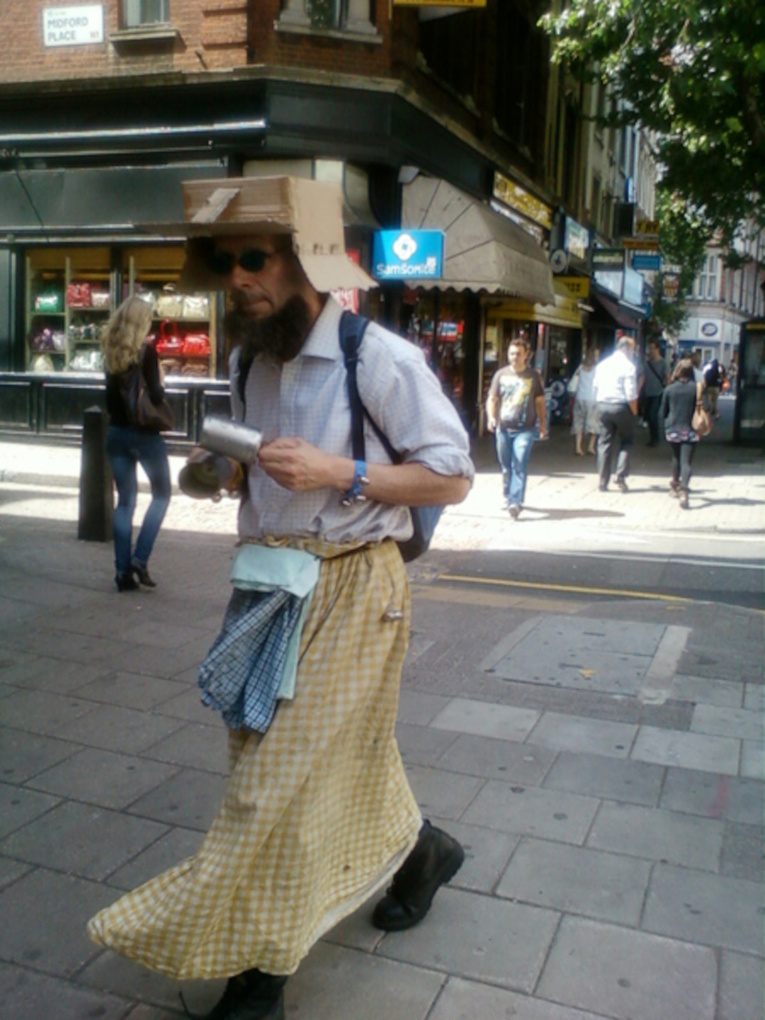 Man wearing a cardboard box hat and a long, yellow chequered skirt. He carries a backpack and what appears to be a tin can in his hands. He is walking down a city street, past shops, and other pedestrians. The overall impression is one of eccentricity and perhaps homelessness or poverty. The man’s attire is unusual and stands out from the more conventional clothing of the people around him. The scene is brightly lit, suggesting it's daytime.