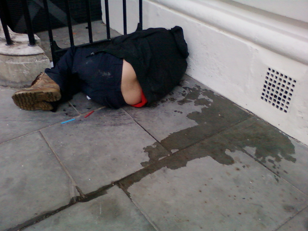 Person lying on the ground next to a building. The individual appears to be slumped over, partially obscuring their face, and wearing dark clothing. A wet patch is visible on the pavement near them. There are also small, possibly discarded, items on the ground. The overall impression is one of someone who may be incapacitated or homeless.