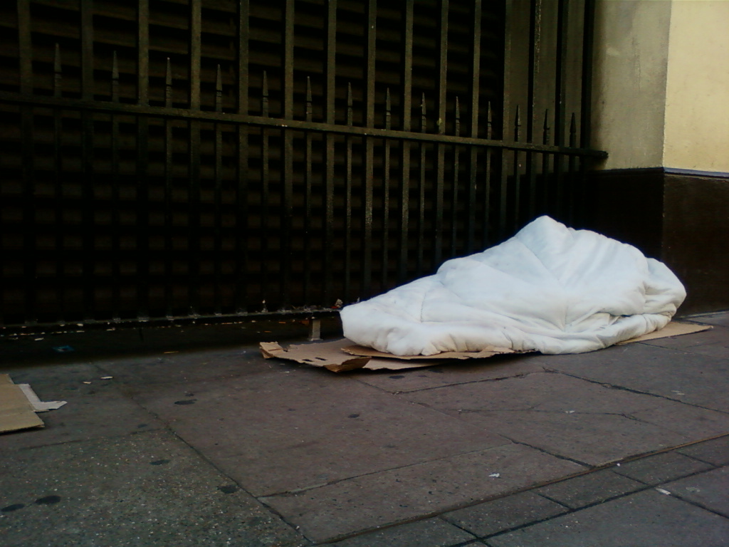 White, puffy comforter or duvet lying on a piece of cardboard on a city side walk next to a dark metal fence. The scene suggests homelessness or destitution.