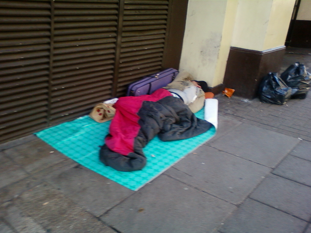 Person sleeping rough on a city side walk. They are curled up in a sleeping bag on a small, teal-coloured mat, with a purple suitcase and some belongings next to them. The setting appears to be an urban area, next to a building with dark brown slatted shutters. There are also some black plastic bags nearby. The overall impression is one of homelessness and poverty.