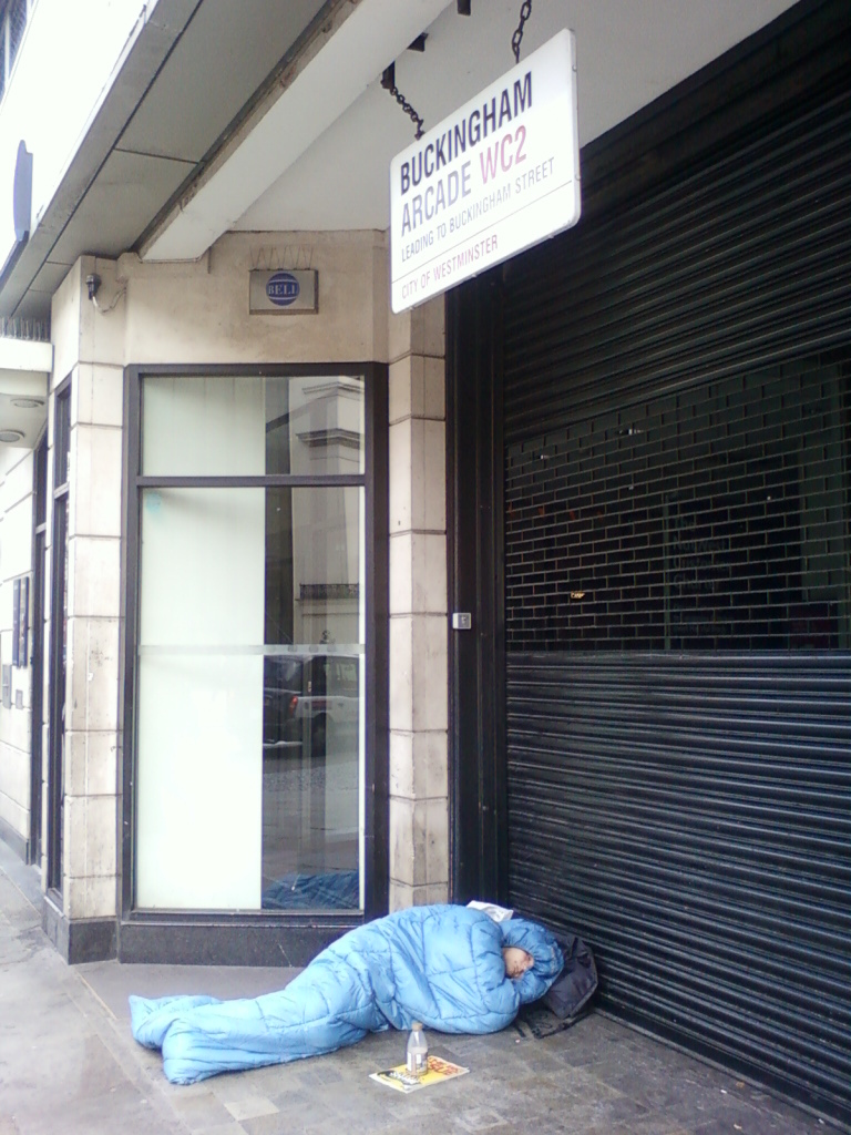 Person sleeping rough in a sleeping bag on the pavement outside Buckingham Arcade in London. The contrast between the person's vulnerable state and the upscale location of the arcade is striking. The image's composition emphasises this juxtaposition, with the building's elegant signage and modern storefront clearly visible above the homeless individual.