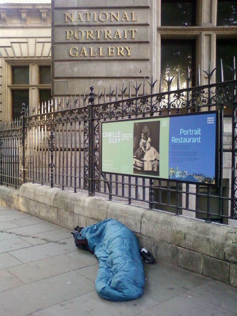 Person sleeping rough in a blue sleeping bag on the pavement outside the National Portrait Gallery in London. A sign advertising the gallery's restaurant and an exhibition on Camille Silvy is visible in the background. The juxtaposition highlights the stark contrast between the cultural institution and the plight of homelessness.