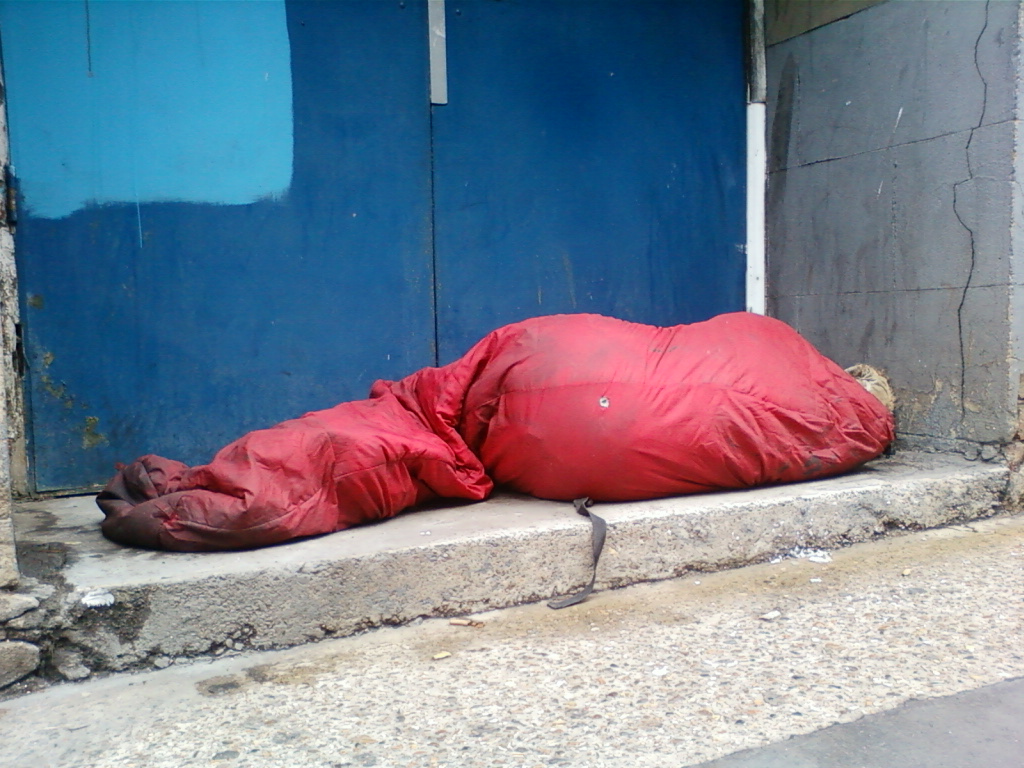 Person curled up and sleeping in a red sleeping bag against a blue door and a grey wall. The setting appears to be outdoors, possibly in an alleyway or some other less-than-ideal location. The overall impression is one of homelessness or hardship.