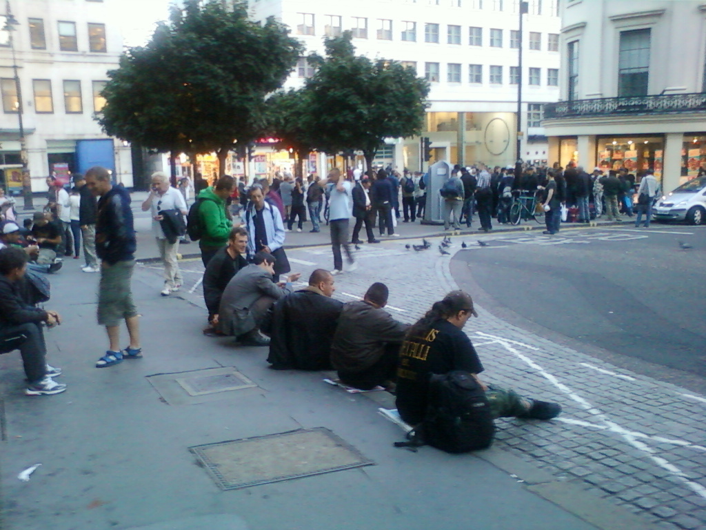 Street scene where a group of young men are sitting on the pavement. They appear to be casually dressed and some are interacting with each other. In the background, a larger crowd of pedestrians are walking along a busy street lined with shops and buildings. The overall atmosphere seems relatively relaxed, though the reason for the group's presence on the pavement is unclear, possibly waiting for something or simply resting. Several pigeons are also visible on the street. The image is somewhat grainy and taken from a distance, suggesting a candid snapshot rather than a staged photograph.
