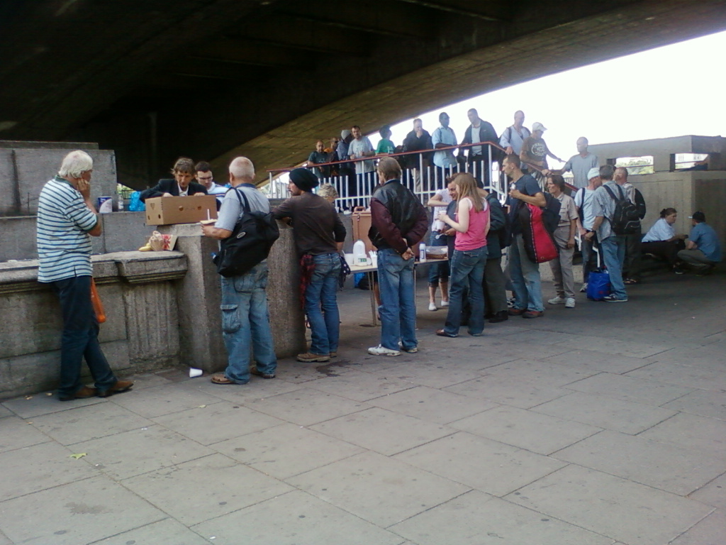 Group of people gathered under a bridge. A smaller group is near a stone structure, seemingly participating in some sort of exchange or transaction – possibly food distribution; a cardboard box is visible. A larger group stands further back, some observing, others seemingly waiting. The setting suggests an outdoor, possibly public space. The overall mood is casual and somewhat informal.