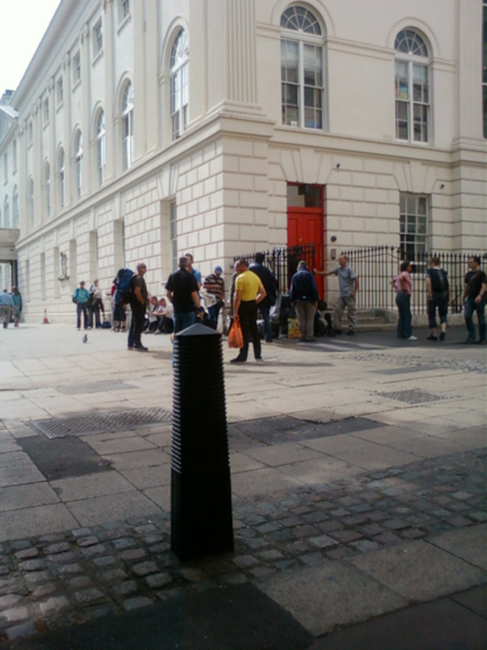 Group of people gathered in front of a large, classical-style building.  The building is light-colored with many evenly spaced windows and a prominent red door. The people appear to be casually dressed, some carrying bags or backpacks. The scene suggests a possibly informal gathering or a moment of pause outside the building. The paving in the foreground is a mix of newer and older materials.