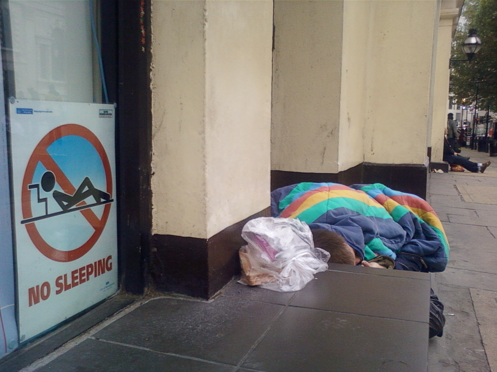 Homeless person sleeping on the ground next to a building. They are wrapped in a colourful blanket. Next to them is a No Sleeping sign. The juxtaposition of the sign and the sleeping person highlights the harsh realities of homelessness and the societal pressures faced by those experiencing it.