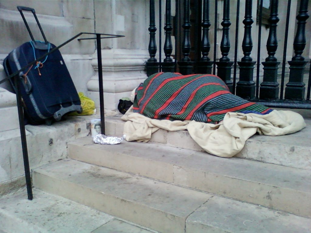 Person sleeping rough on some stone steps outside a building. They are covered with blankets and a striped throw. A suitcase and some other belongings are next to them. The scene suggests homelessness and hardship.