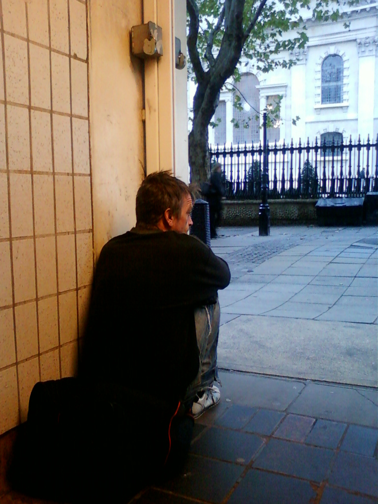 A man, seemingly homeless, sitting hunched over on the ground in a doorway. He's wearing dark clothing and jeans, and a bag rests beside him. The background shows a neatly kept, affluent-looking street with a fence and elegant buildings. The contrast between the man’s impoverished state and the backdrop of wealth is striking. The man appears withdrawn and contemplative.