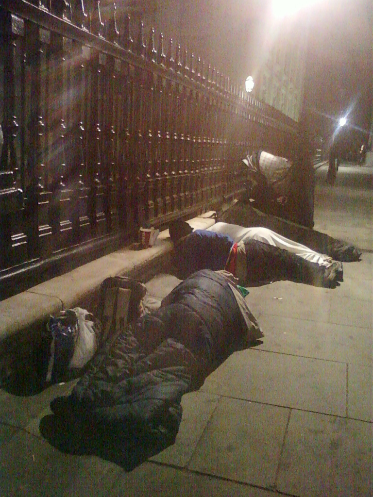 Two individuals sleeping rough on a city pavement at night. They are wrapped in sleeping bags near a tall, dark fence, suggesting a location possibly near a building or park. The scene is dimly lit, emphasising the vulnerability and isolation of the individuals. The overall impression is one of poverty and homelessness.