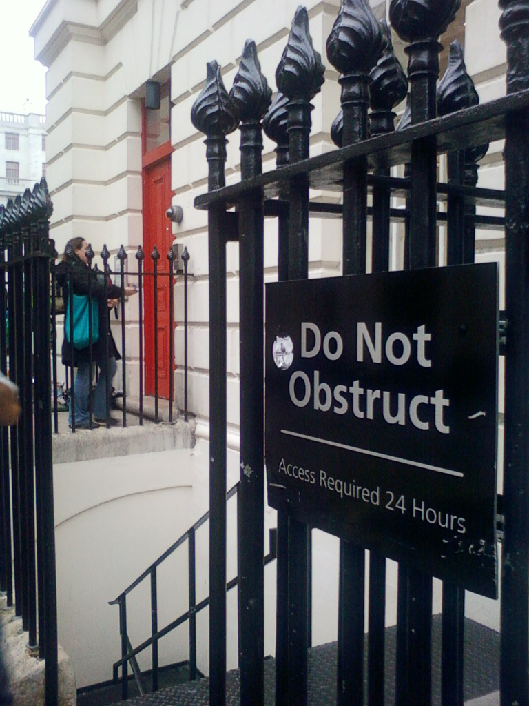 Woman using a doorbell at a red door. The door is behind a black metal fence with finials.  A sign on the fence reads Do Not Obstruct - Access Required 24 Hours. The setting appears to be a building in a city. The overall impression is one of a somewhat utilitarian and possibly official or institutional setting.