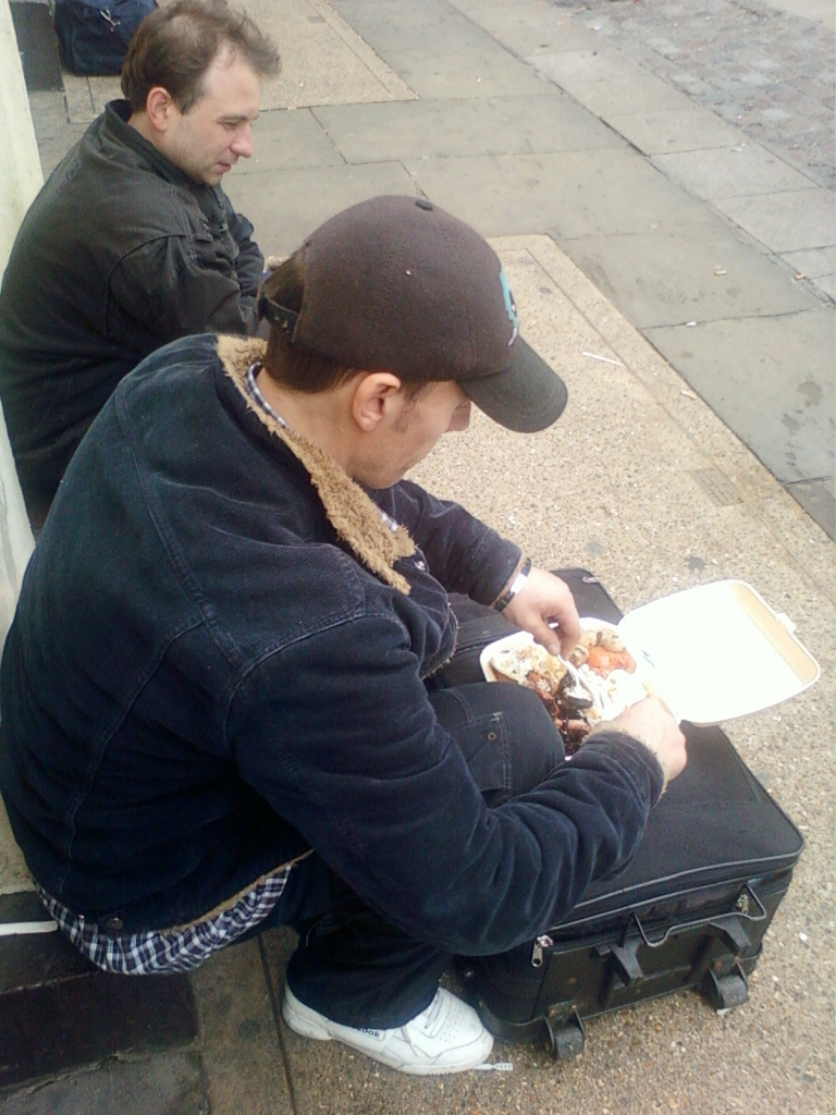 Two men sitting on a sidewalk, one of whom is eating from a takeout container. The man eating is wearing a dark jacket and cap, and is seated on a suitcase. The other man is sitting beside him, appearing somewhat withdrawn. The overall setting suggests a street scene, possibly in a city. The mood is somewhat somber and possibly reflects a situation of hardship or transient living.