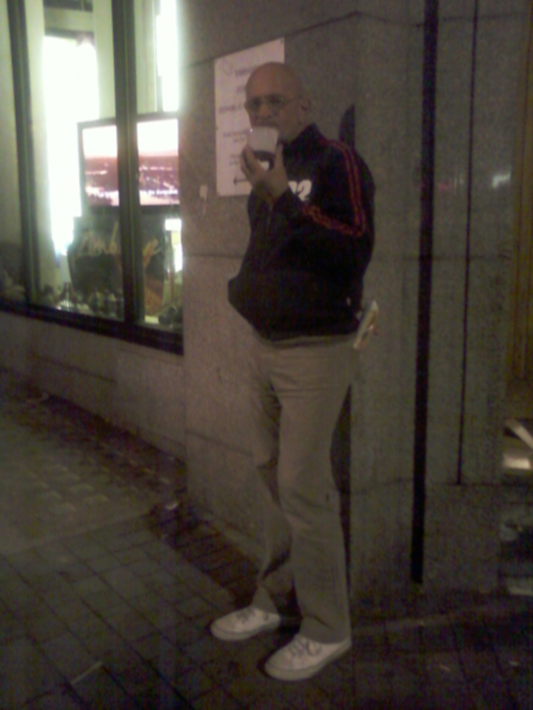 Bald, light-skinned man standing outside at night, leaning against a stone building. He is wearing a dark jacket with red stripes on the sleeves, light beige pants, and white sneakers. He appears to be holding and drinking from a small white cup or container. The background includes a storefront with windows displaying interior items and screens showing images, possibly from a shop or business. The overall ambience is dimly lit and somewhat sombre.