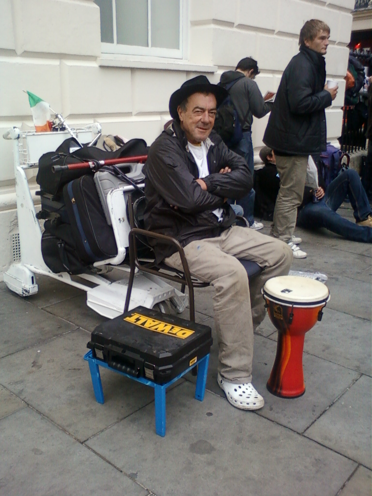 Man sitting in a chair attached to a motorised luggage cart. He is wearing a black jacket, khaki pants, and a hat. He has his arms crossed and appears relaxed. He has a small djembe drum next to him and a DeWalt toolbox on a blue stool. In the background, there are other people, and an Irish flag is visible on the luggage cart. The setting appears to be an urban location, possibly a city street. The overall impression is of a street musician or performer who is comfortably settled in for a performance or break.