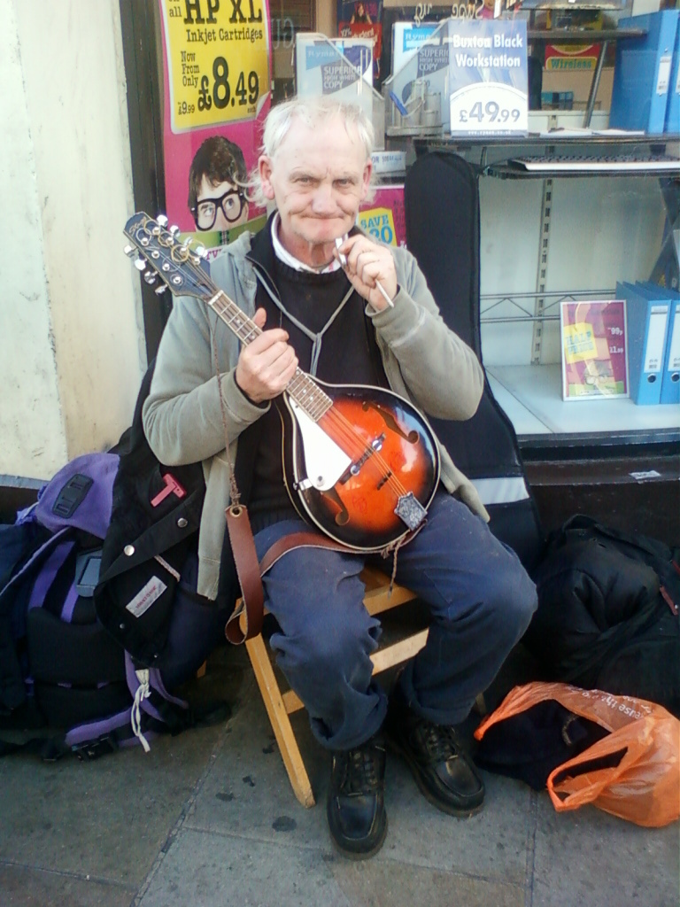 Middle-aged, light-skinned man with short, light blonde hair sitting on a small wooden stool outside a shop. He is playing a mandolin, holding it gently in his hands. He’s wearing a light grey or beige jacket, a dark colored shirt underneath, dark blue jeans, and dark brown work boots. He appears to be casually dressed, and his expression is somewhat pensive or thoughtful. He is holding something small (possibly a toothpick or cigarette) near his mouth with his right hand. The setting is urban, with a shop window in the background filled with various items including ink cartridges, a computer workstation, and office supplies. There are posters advertising discounted prices. Several backpacks are on the ground beside the musician suggesting he may be a street performer or traveler. An orange plastic bag lies on the ground near his feet. The overall mood is somewhat quiet and observational. 